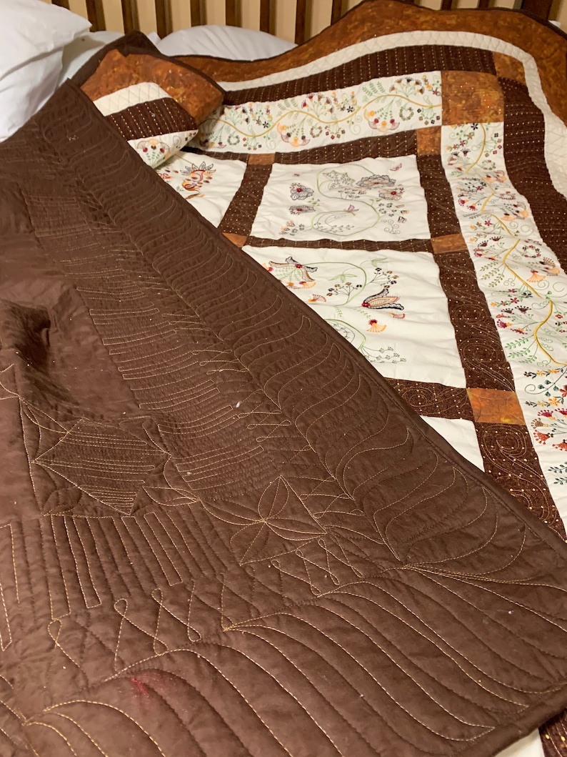 Puede incluir: Una manta acolchada marr&oacute;n con un patr&oacute;n decorativo de costuras. La manta est&aacute; doblada sobre una colcha blanca y marr&oacute;n con dise&ntilde;os florales bordados.
