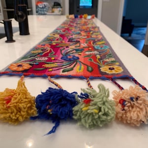 May include: A colorful table runner with embroidered flowers and tassels. The runner is made of wool and has a blue, red, pink, yellow, and green color scheme.