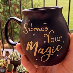 May include: Black ceramic mug with a gold inscription that reads "Embrace Your Magic" with small gold stars scattered around the text.