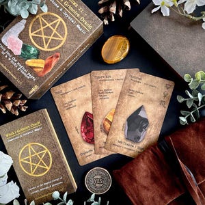 May include: A collection of oracle cards, a book, and a box with a pentagram design. The cards feature gemstone illustrations and text. The box and book are brown with gold lettering. A tiger's eye stone and a coin with the word "YES" are also present.
