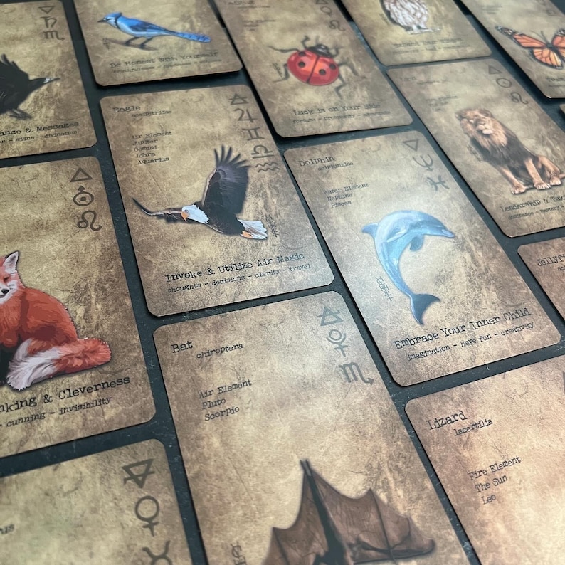 May include: A collection of tarot cards with vintage-style illustrations of animals and symbols. Each card features a different animal, such as a blue jay, eagle, and dolphin, with accompanying text and astrological symbols. The cards have a distressed, aged appearance.