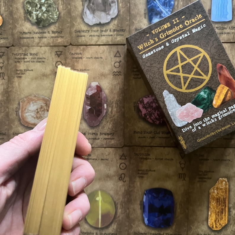May include: A deck of gold-edged cards is held in front of a box labeled "VOLUME II - Witch's Grimoire Oracle Gemstone & Crystal Magic." The box features a pentagram and illustrations of gemstones. The background includes cards with gemstone names.