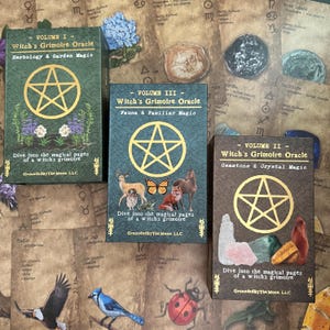 May include: Three oracle card decks titled "Witch's Grimoire Oracle" with different volumes: Herbology & Garden Magic, Fauna & Familiar Magic, and Gemstone & Crystal Magic. Each deck features a gold pentagram design and the text "Dive into the magical pages of a witch's grimoire."