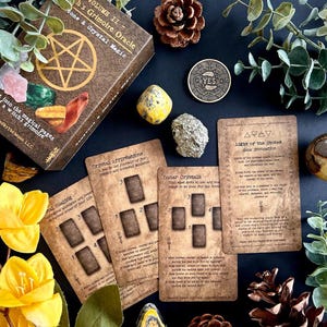 May include: A collection of spiritual items including a brown box with text, crystals, cards, and a coin with "YES" on it. The box reads "Volume II - Witch's Grimoire Oracle". The cards have text and diagrams. The scene is decorated with flowers and greenery.