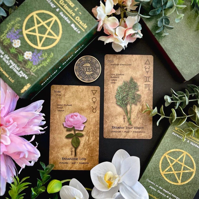 May include: An arrangement featuring a green box set, oracle cards, and a coin with the word "YES". The cards display botanical illustrations and text. Pink and white flowers and greenery surround the items, creating a mystical theme.