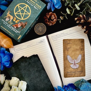 May include: A brown and white tarot card with the word "Dove" and the symbol of the scales. The card has a white dove with outstretched wings on a brown background. The text "Inner Peace & Harmony" is at the bottom of the card. The card is on a black surface with a green journal and a deck of tarot cards.