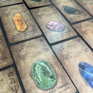 May include: A collection of oracle cards with various gemstones and text. The cards have a vintage, textured appearance with symbols and phrases like "Rekindle Your Energy" and "Mend Your Old Wounds". The gemstones include a green serpentine and a pink stone.