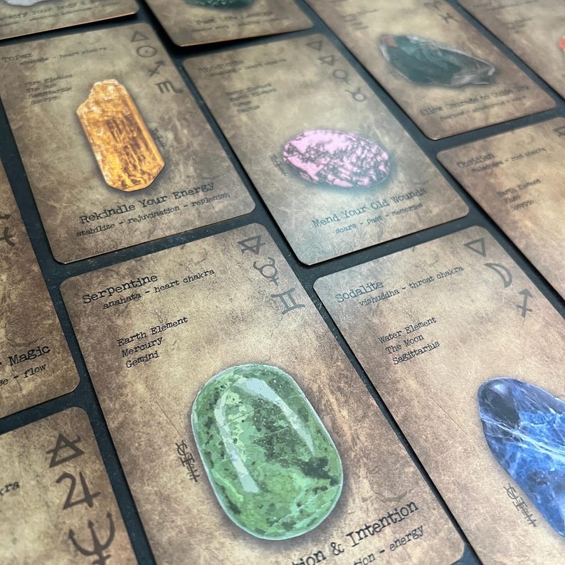 May include: A collection of oracle cards with various gemstones and text. The cards have a vintage, textured appearance with symbols and phrases like "Rekindle Your Energy" and "Mend Your Old Wounds". The gemstones include a green serpentine and a pink stone.