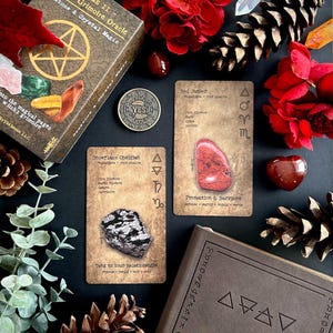 May include: A still-life arrangement featuring a grimoire oracle set. The set includes a box, cards with text, a coin, and crystals. The cards display images of crystals and text. Red flowers and pinecones add decorative elements.