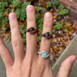 May include: Three wire-wrapped rings with different colored gemstones. The rings are on a person's hand. The ring on the index finger has a red gemstone, the ring on the middle finger has a purple gemstone, and the ring on the ring finger has a blue gemstone.