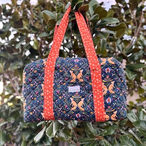May include: A quilted tote bag with a dark blue floral and fox pattern. The bag has bright orange handles with a white polka dot design. The bag is hanging from a tree branch.