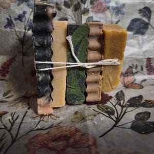 May include: A stack of five rectangular soap bars tied together with white twine. The bars feature varied textures and colours, including black and white marbled, solid beige, dark green with black patterns, ridged brown, and solid mustard yellow.