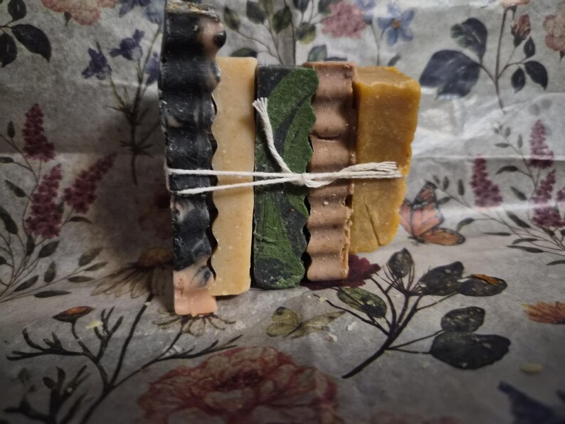 May include: A stack of five rectangular soap bars tied together with white twine. The bars feature varied textures and colours, including black and white marbled, solid beige, dark green with black patterns, ridged brown, and solid mustard yellow.
