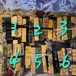May include: Six bundles of rectangular soap bars, each secured with a thin black or white elastic band. The bars feature varied textures and colors, including shades of green, yellow, brown, and cream. Each bundle is numbered with a cyan digit.