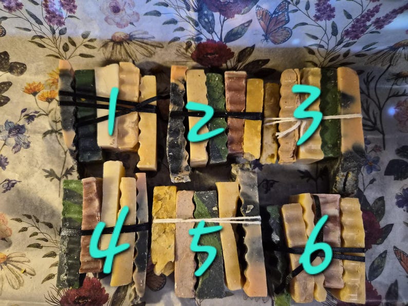 May include: Six bundles of rectangular soap bars, each secured with a thin black or white elastic band. The bars feature varied textures and colours, including shades of green, yellow, brown, and cream. Each bundle is numbered with a cyan digit.