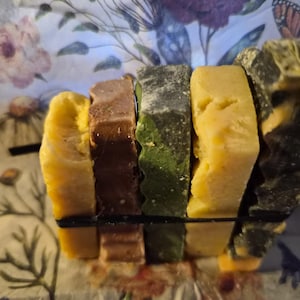 May include: A stack of five rectangular bars of soap held together by a thin black elastic band. The bars feature varied colours including yellow, brown, dark green, light yellow, and a marbled black and yellow pattern with a textured surface.