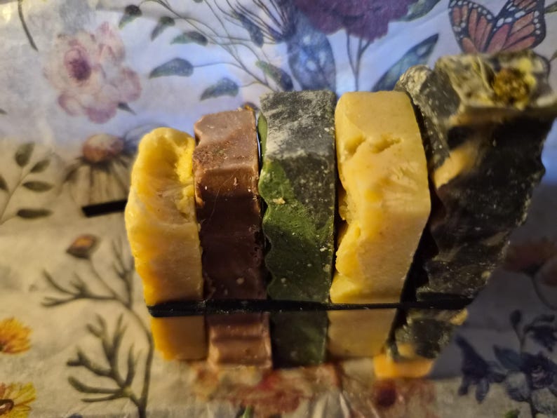 May include: A stack of five rectangular bars of soap held together by a thin black elastic band. The bars feature varied colours including yellow, brown, dark green, light yellow, and a marbled black and yellow pattern with a textured surface.