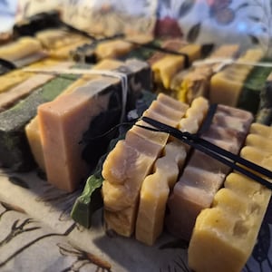 May include: A collection of rectangular soap bars bundled together with thin black and white elastic bands. The bars feature various colours including shades of yellow, green, brown, and cream, with some having smooth surfaces and others showing wavy edges.
