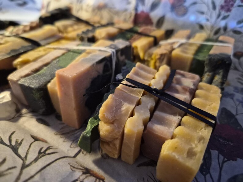 May include: A collection of rectangular soap bars bundled together with thin black and white elastic bands. The bars feature various colours including shades of yellow, green, brown, and cream, with some having smooth surfaces and others showing wavy edges.