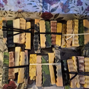 May include: Six bundles of rectangular soap bars, each tied with black or white string. The bars feature varied textures and colours including shades of green, yellow, brown, cream, and grey, arranged in stacks of four or five pieces per bundle.