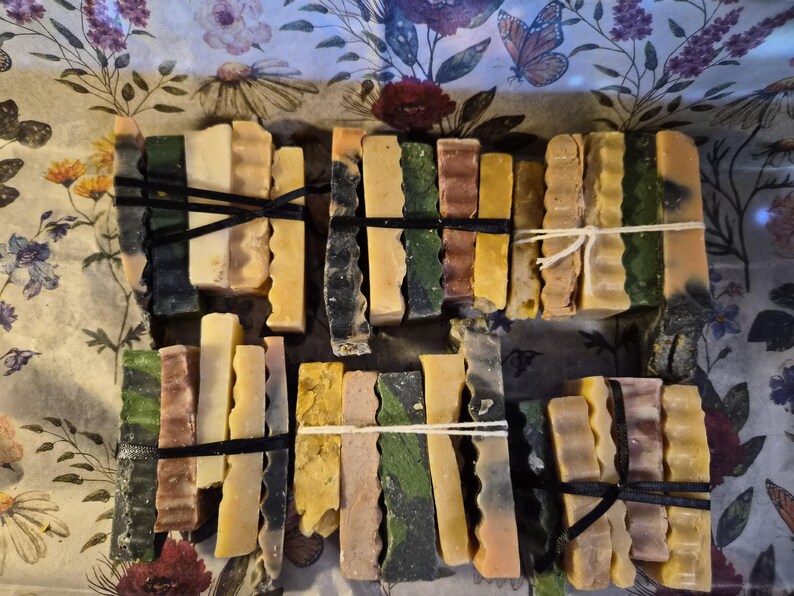 May include: Six bundles of rectangular soap bars, each tied with black or white string. The bars feature varied textures and colours including shades of green, yellow, brown, cream, and grey, arranged in stacks of four or five pieces per bundle.