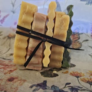 May include: A stack of four rectangular soap bars tied together with a thin black ribbon. The bars feature varied textures, including smooth and wavy edges, in shades of yellow, light brown, and dark green, arranged vertically in a bundle.