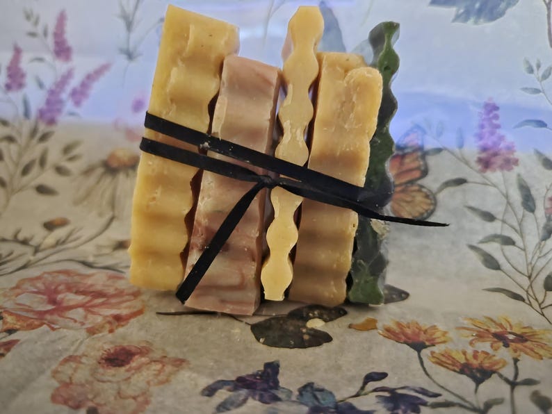 May include: A stack of four rectangular soap bars tied together with a thin black ribbon. The bars feature varied textures, including smooth and wavy edges, in shades of yellow, light brown, and dark green, arranged vertically in a bundle.