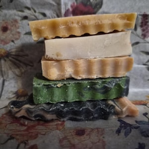 May include: A vertical stack of five rectangular bars of soap. The top bar is light tan with a wavy edge, followed by a smooth cream bar, a tan bar with a wavy edge, a dark green bar with a textured edge, and a bottom bar with a wavy black and tan swirl.