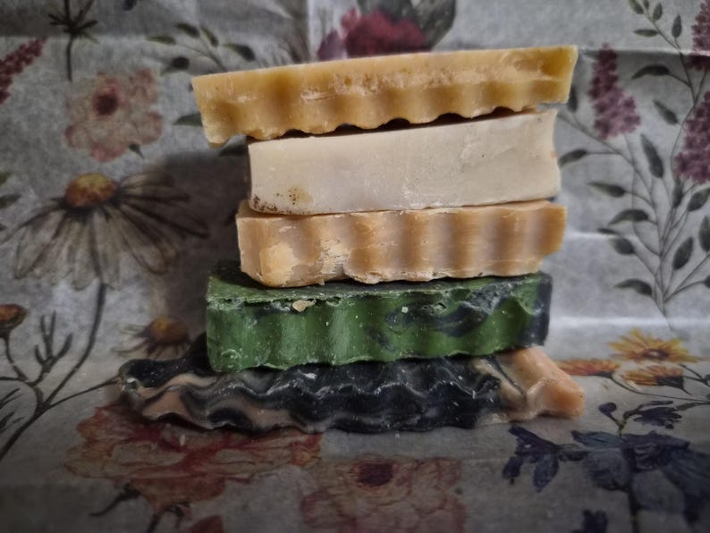 May include: A vertical stack of five rectangular bars of soap. The top bar is light tan with a wavy edge, followed by a smooth cream bar, a tan bar with a wavy edge, a dark green bar with a textured edge, and a bottom bar with a wavy black and tan swirl.