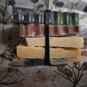 May include: A stack of five rectangular bars of soap in varying colours including dark green, mauve, cream, yellow, and a marbled grey-orange, all secured together by a single vertical black ribbon wrapped around the centre of the bars.