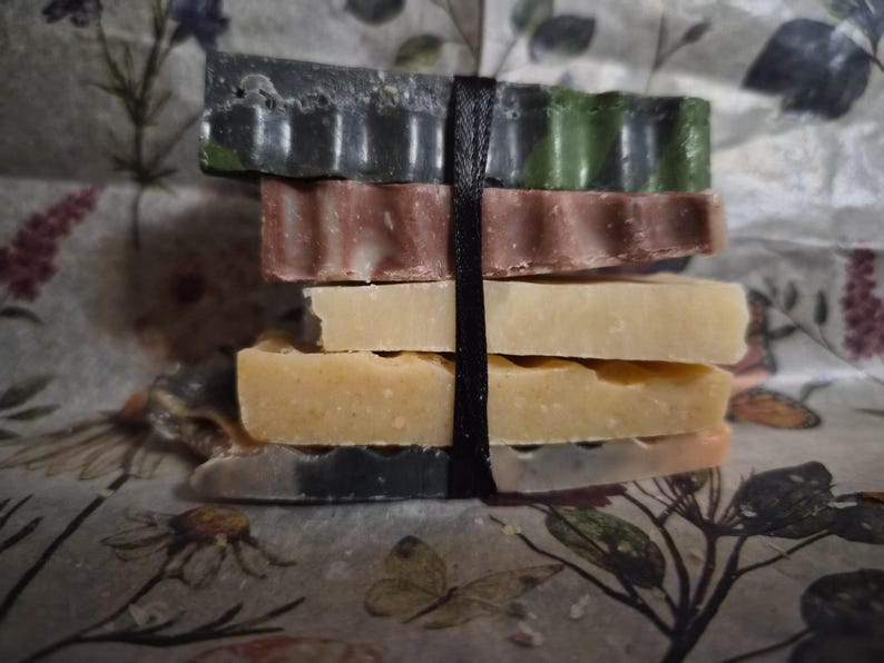 May include: A stack of five rectangular bars of soap in varying colours including dark green, mauve, cream, yellow, and a marbled grey-orange, all secured together by a single vertical black ribbon wrapped around the centre of the bars.