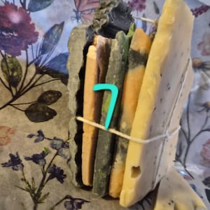 May include: A stack of rectangular soap bars in varying shades of beige, green, and orange, secured together with a thin white string. A bright turquoise number 7 is placed on the front of the stack. The soap bars have textured edges.