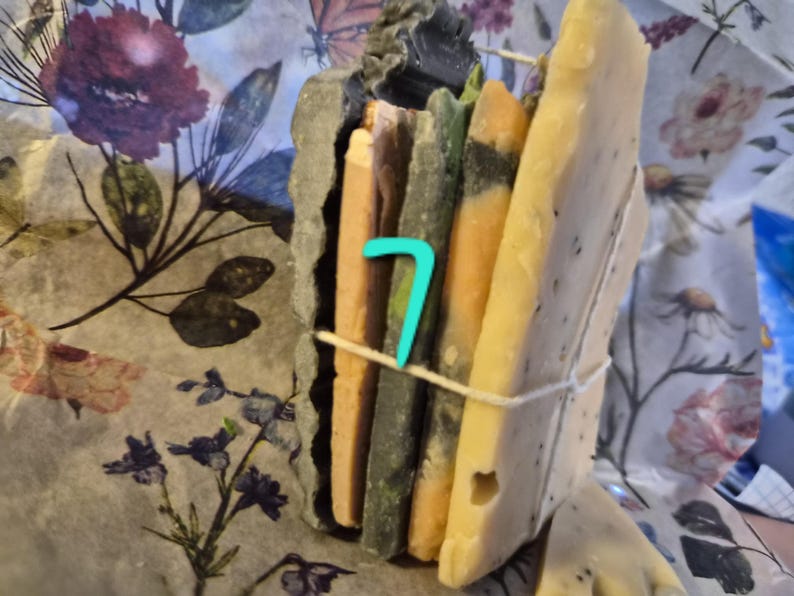 May include: A stack of rectangular soap bars in varying shades of beige, green, and orange, secured together with a thin white string. A bright turquoise number 7 is placed on the front of the stack. The soap bars have textured edges.