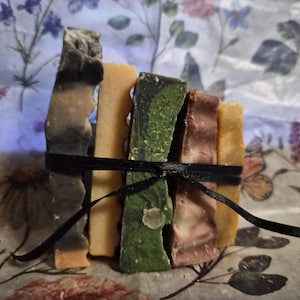 May include: A stack of five rectangular bars of soap tied together with a thin black ribbon. The bars feature varied colours including charcoal grey, solid light orange, marbled forest green, muted mauve, and a pale yellow, all with textured edges.
