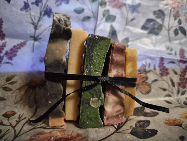 May include: A stack of five rectangular bars of soap tied together with a thin black ribbon. The bars feature varied colours including charcoal grey, solid light orange, marbled forest green, muted mauve, and a pale yellow, all with textured edges.