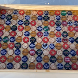 May include: A wooden tray with a mosaic of bottle caps, featuring a variety of brands including Corona Extra, IBC Root Beer, and King Cobra. The caps are arranged in a grid pattern and are set in a clear resin.