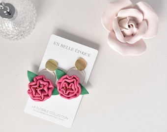 Op de afbeelding: Een paar roze bloemoorbellen met gouden accenten. De oorbellen liggen op een witte kaart met de tekst "En Belle Epoque" en "La Florale Collection" in zwart.