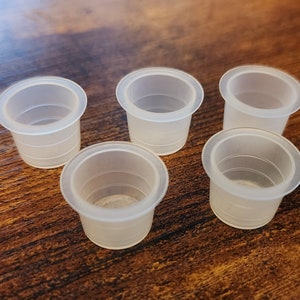 Könnte beinhalten: Fünf transparente Plastikbecher, wahrscheinlich für Tattootinte, auf einer Holzoberfläche angeordnet.