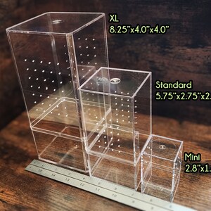 以下が含まれることがあります： XL、スタンダード、ミニの3つの異なるサイズの透明アクリルボックス。ボックスは、一番大きいボックスが下、一番小さいボックスが上に積み重ねられています。ボックスは、各側面に小さな穴のグリッドがあります。ボックスは木製の表面の上にあります。各ボックスの寸法は、ラベルの下に記載されています。
