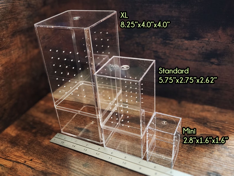 以下が含まれることがあります： XL、スタンダード、ミニの3つの異なるサイズの透明アクリルボックス。ボックスは、一番大きいボックスが下、一番小さいボックスが上に積み重ねられています。ボックスは、各側面に小さな穴のグリッドがあります。ボックスは木製の表面の上にあります。各ボックスの寸法は、ラベルの下に記載されています。
