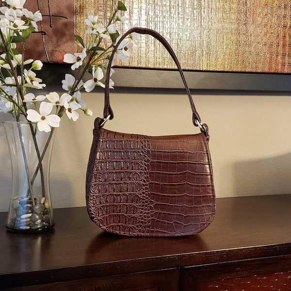 May include: A brown handbag with a crocodile skin pattern. The bag has a curved shape, a short handle, and silver-colored hardware. The bag is sitting on a dark brown surface.