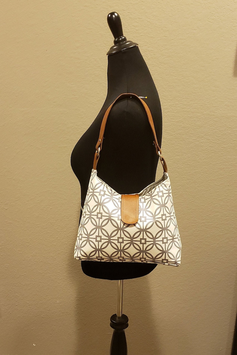 May include: A white and gray geometric patterned handbag with a brown leather strap and handle.