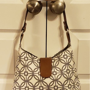 May include: A white and gray geometric patterned handbag with a brown leather strap and handle. The bag is hanging on two door knobs.