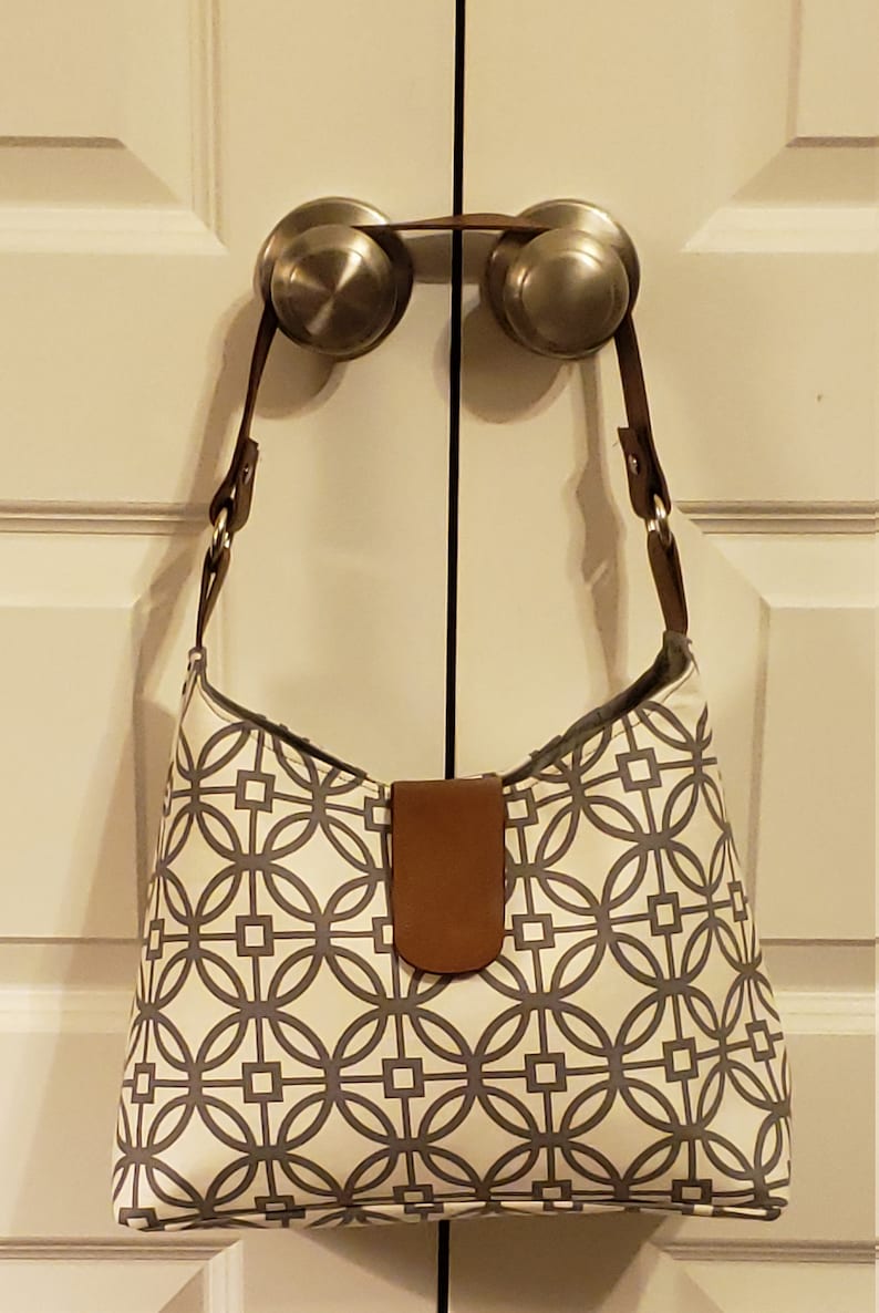 May include: A white and gray geometric patterned handbag with a brown leather strap and handle. The bag is hanging on two door knobs.