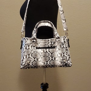 May include: A black and white snakeskin-patterned handbag with a top handle and a shoulder strap. The bag is displayed on a black tripod stand.
