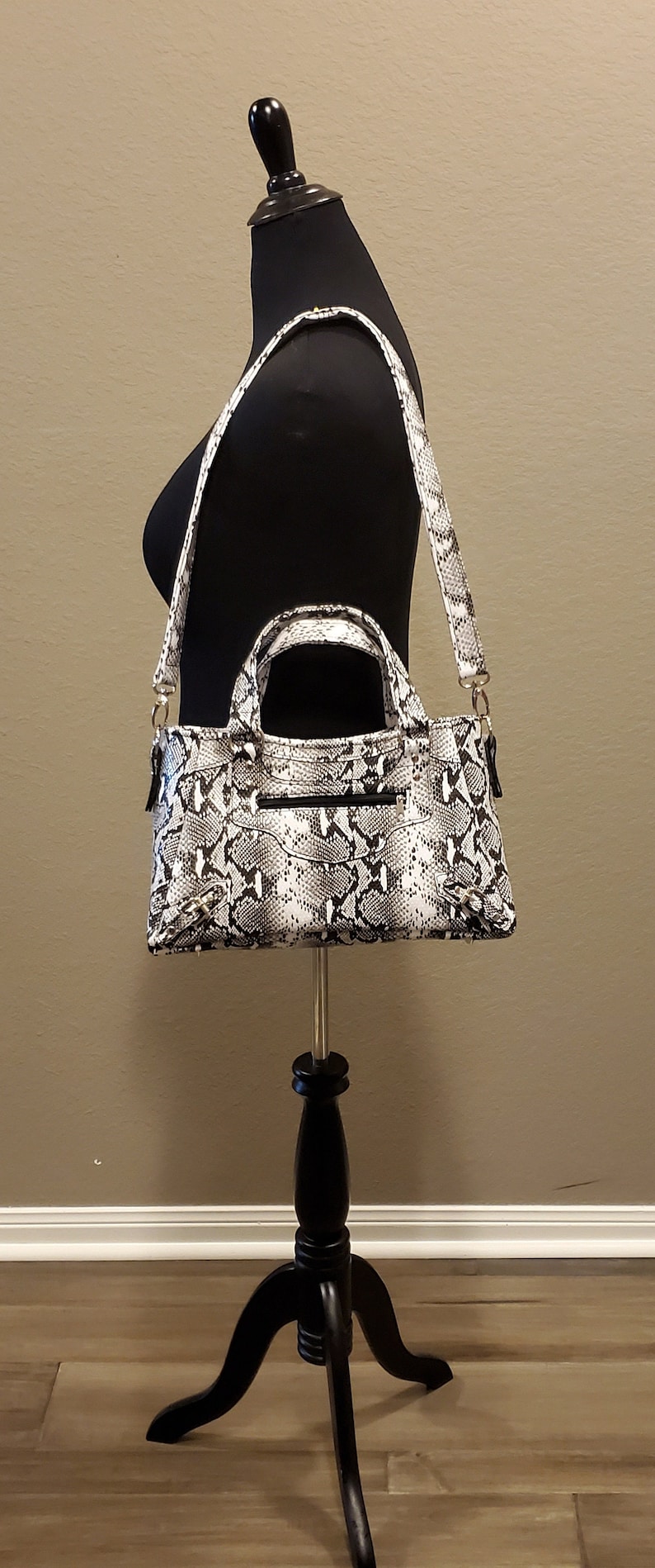 May include: A black and white snakeskin-patterned handbag with a top handle and a shoulder strap. The bag is displayed on a black tripod stand.