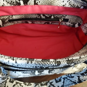 May include: A black and white snakeskin print handbag with a pink fabric lining and a zippered pocket. The bag has a top handle and a zipper closure.