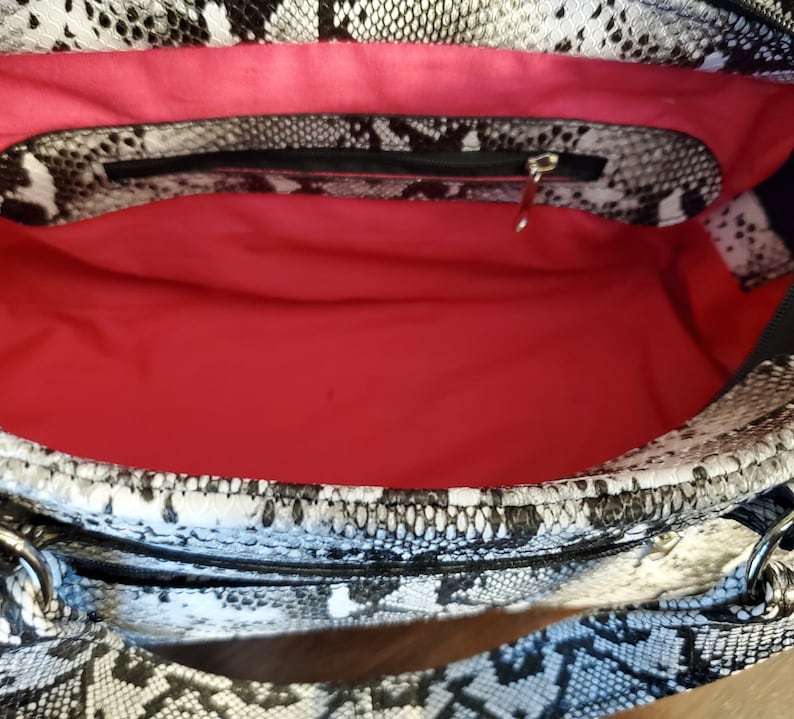 May include: A black and white snakeskin print handbag with a pink fabric lining and a zippered pocket. The bag has a top handle and a zipper closure.