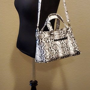 May include: A black and white snakeskin handbag with a gold chain strap hanging on a black mannequin. The handbag has a top handle and a zipper closure.