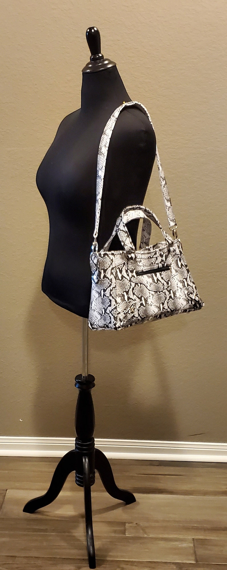 May include: A black and white snakeskin handbag with a gold chain strap hanging on a black mannequin. The handbag has a top handle and a zipper closure.